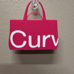 May include: A bright pink, textured, flat, handbag-shaped object with two handles. The word "Curv" is printed in large, bold, white letters across the front. The object is hanging from a clear plastic hook attached to a textured wall surface.