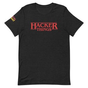 May include: Dark gray t-shirt with the red text "HACKER THINGS" across the chest. The text is in a bold, retro font. The left sleeve has the text "CAL EAT" in yellow. The shirt is made of a soft material.