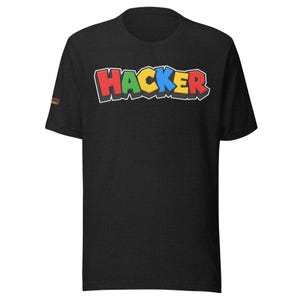 May include: A black short-sleeve t-shirt featuring the word HACKER printed across the chest in a multi-colored, blocky, retro-style font with red, green, yellow, and blue letters. A small logo is visible on the right sleeve of the garment.