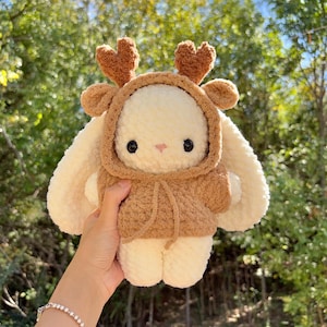 May include: A handmade plush bunny wearing a brown hooded sweater with deer antlers. The bunny is cream-colored with black eyes and a pink nose. The plushie is soft and cuddly, perfect for a child's toy or nursery decor.