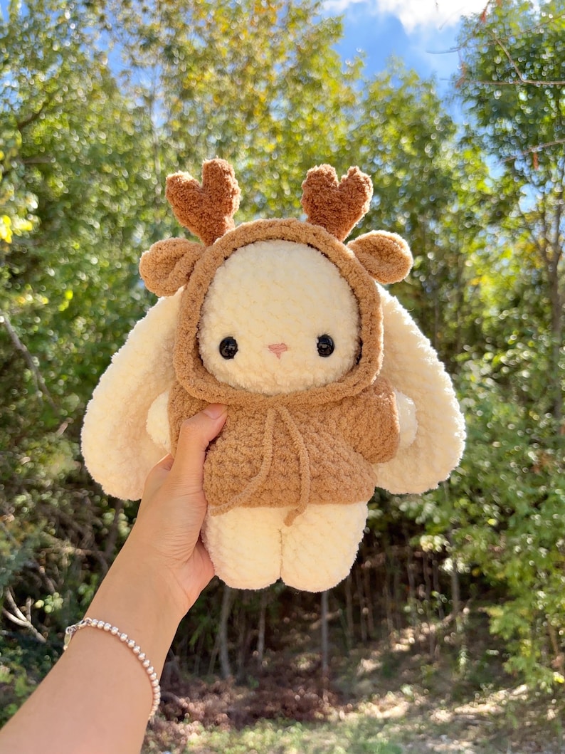 May include: A handmade plush bunny wearing a brown hooded sweater with deer antlers. The bunny is cream-colored with black eyes and a pink nose. The plushie is soft and cuddly, perfect for a child's toy or nursery decor.