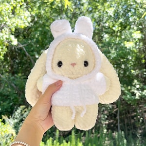 May include: A plush, cream-colored bunny wearing a white hooded sweatshirt. The bunny has large, floppy ears, black eyes, and a small pink nose. The background is a blurred view of green foliage.