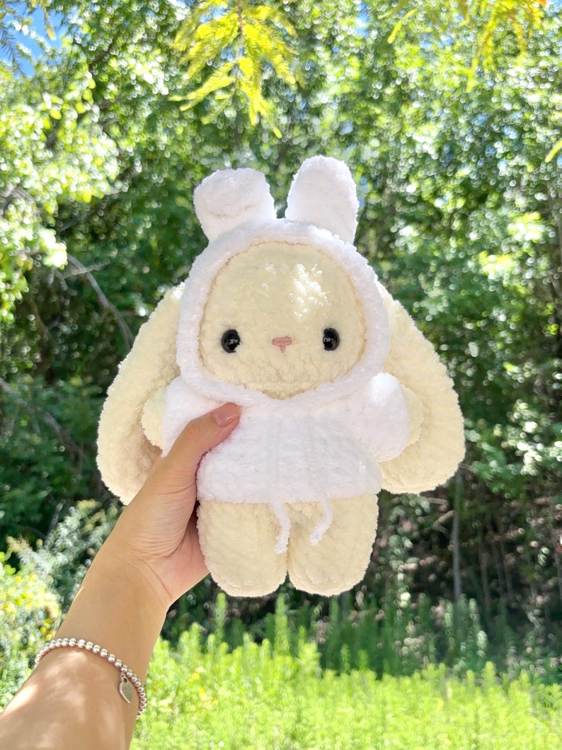 May include: A plush, cream-colored bunny wearing a white hooded sweatshirt. The bunny has large, floppy ears, black eyes, and a small pink nose. The background is a blurred view of green foliage.