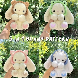 May include: A collection of four plush bunny toys made of soft white yarn, each wearing a different removable hood: brown bear, green frog, pink cat, and blue shark. The text 5 IN 1 BUNNY PATTERN is centered over the images of these stuffed animals.