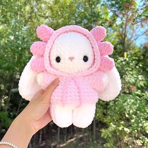 May include: A handmade white and pink plush toy in the shape of a bunny wearing a pink hooded outfit. The bunny has black eyes and a pink nose. The plush toy is held up against a background of green trees.