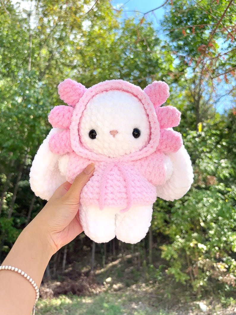 May include: A handmade white and pink plush toy in the shape of a bunny wearing a pink hooded outfit. The bunny has black eyes and a pink nose. The plush toy is held up against a background of green trees.
