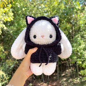 May include: A handmade white plush bunny wearing a black hooded sweatshirt with pink inner ears. The bunny has black button eyes and a pink nose. The plushie is held up against a blurred green background of trees.