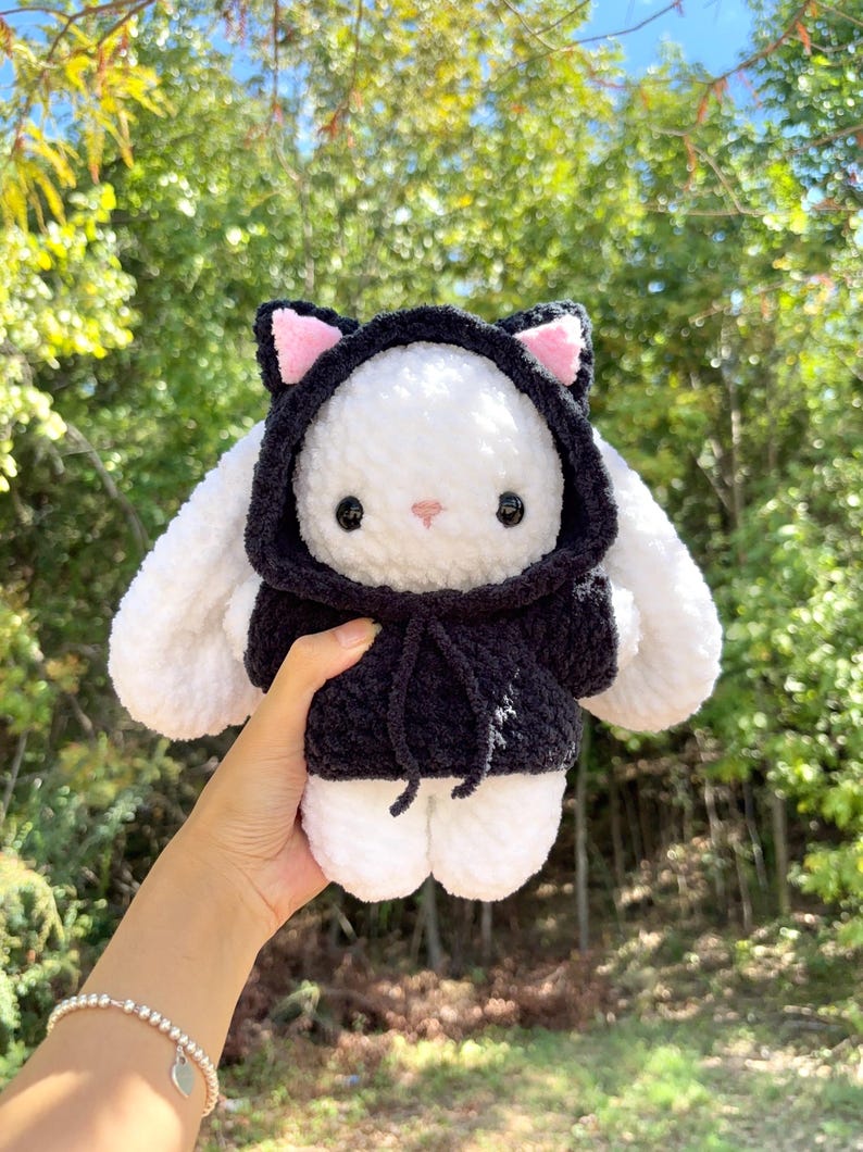 May include: A handmade white plush bunny wearing a black hooded sweatshirt with pink inner ears. The bunny has black button eyes and a pink nose. The plushie is held up against a blurred green background of trees.