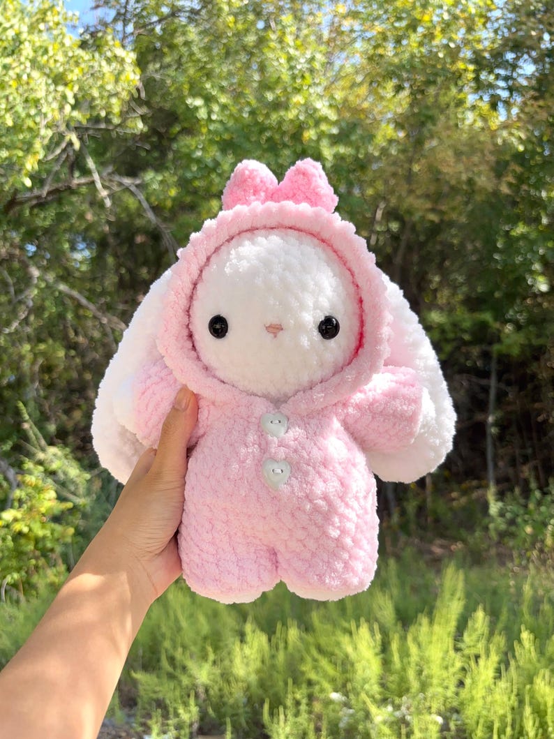 May include: A handmade plush bunny in a pink hooded jumpsuit with a bow on the hood. The bunny has black eyes and a small pink nose. The plushie is held against a backdrop of green foliage.