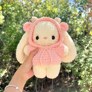 May include: A handmade, plush bunny doll with a pink hooded jacket. The bunny has long, floppy ears and a sweet, embroidered face with black eyes. The doll is cream-colored with pink accents and is soft and cuddly.