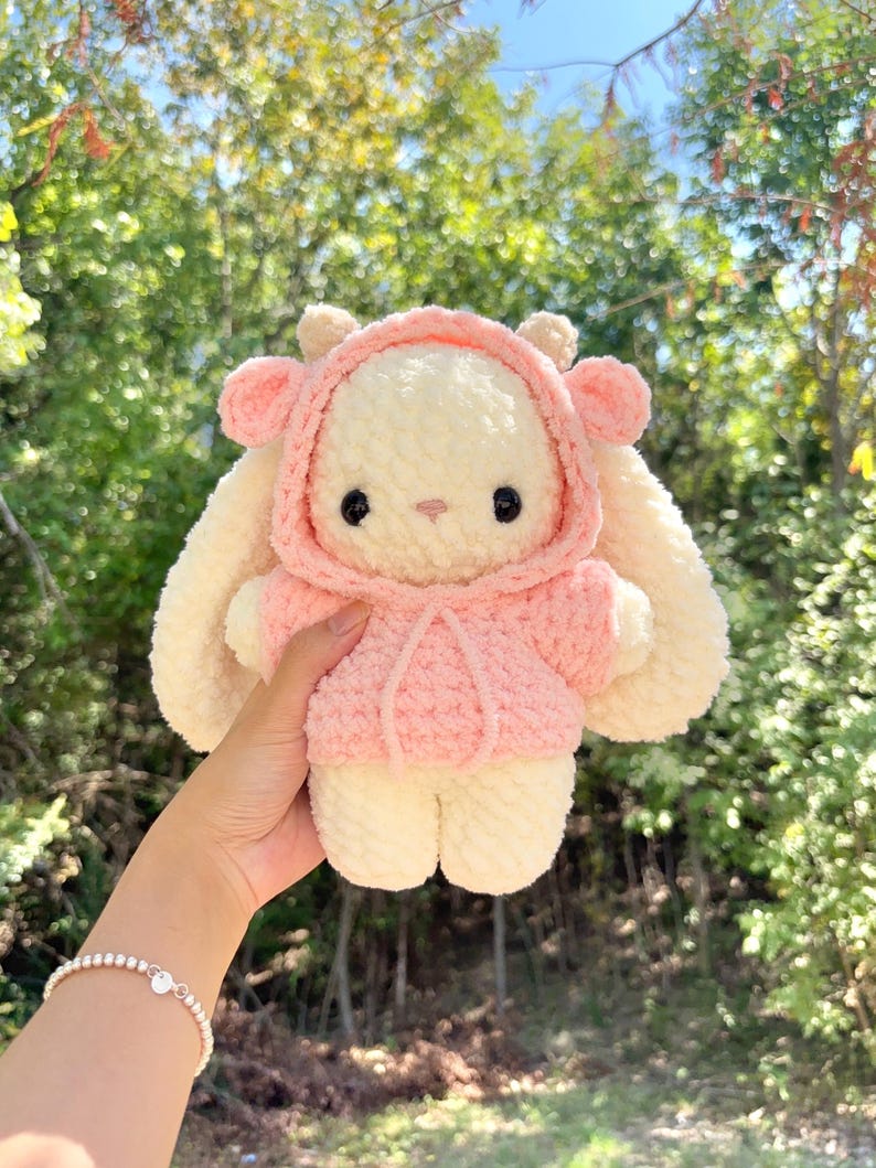 May include: A handmade, plush bunny doll with a pink hooded jacket. The bunny has long, floppy ears and a sweet, embroidered face with black eyes. The doll is cream-colored with pink accents and is soft and cuddly.