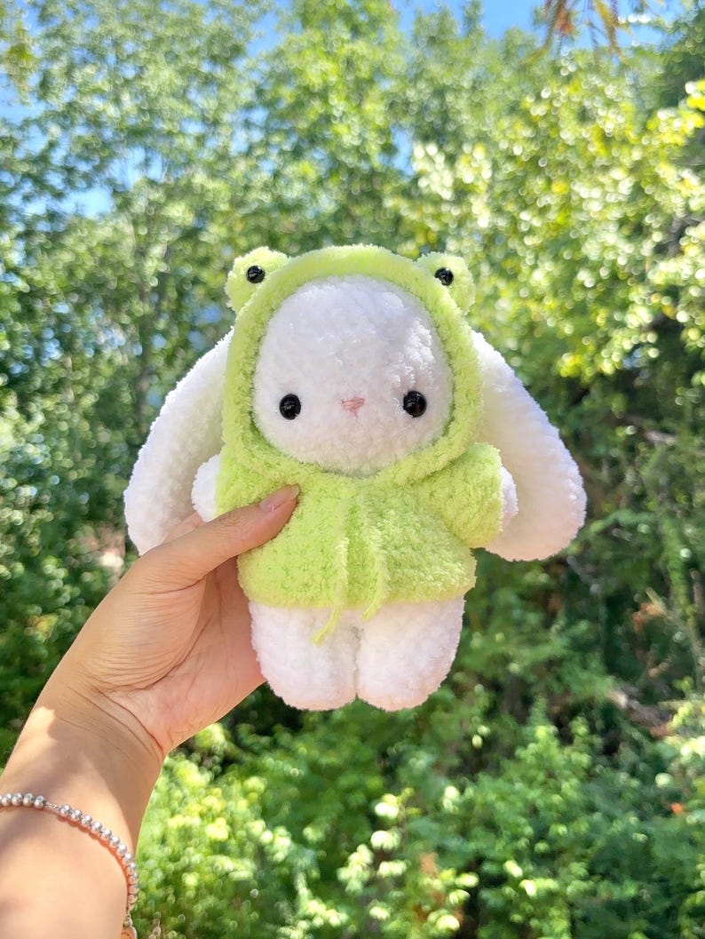 May include: A white plush bunny wearing a light green frog hoodie with black eyes and a pink nose. The bunny's ears and feet are white. The background is a blurred view of green foliage.