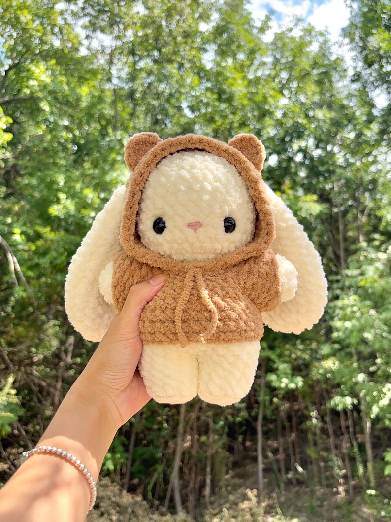 May include: A handmade, plush bunny wearing a brown hooded jacket. The bunny has cream-colored fur, large floppy ears, and black button eyes. The background is a blurred view of green trees.
