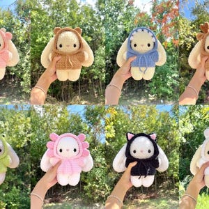 May include: A collection of eight handmade crochet bunny dolls, each wearing a different hooded outfit. The outfits include a pink bear, brown bear, blue shark, brown deer, green frog, pink flower, black cat, and white bunny.