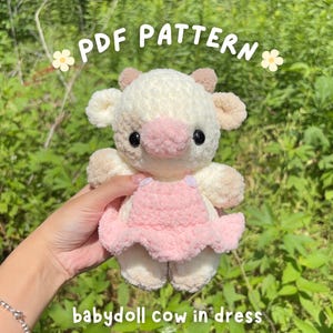 May include: A plush, cream-colored cow with a pink nose and a pink babydoll dress. The cow has small, tan ears and black eyes. Text on the image reads "PDF PATTERN" and "babydoll cow in dress". The background is blurred green foliage.