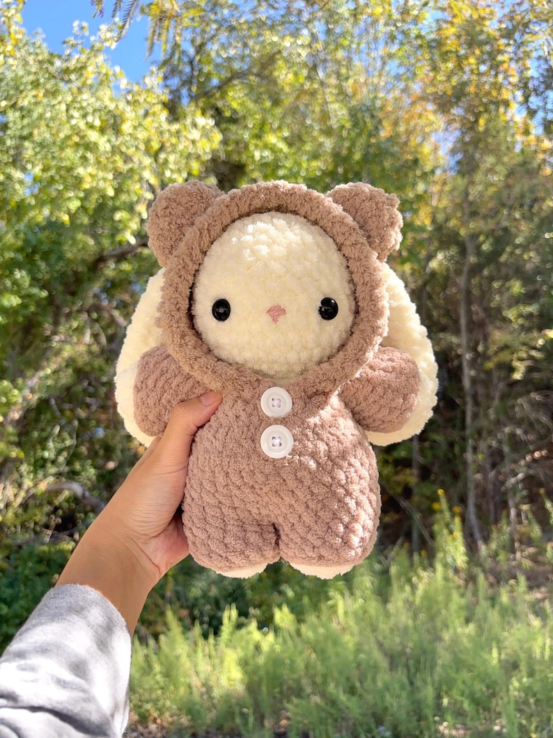 May include: A handmade plush bunny wearing a brown bear costume. The bunny has a cream-coloured body, black eyes, and a pink nose. The bear costume features a hood with bear ears and two white buttons. The plush toy is soft and cuddly.