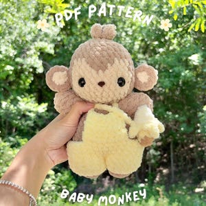 May include: A plush baby monkey toy made of soft brown and tan yarn, wearing yellow overalls and holding a small yellow banana. The text PDF PATTERN and BABY MONKEY is printed in white on the image, which features a textured, fuzzy appearance.