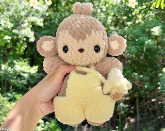 May include: A plush baby monkey toy made of soft brown and tan yarn, wearing yellow overalls and holding a small yellow banana. The text PDF PATTERN and BABY MONKEY is printed in white on the image, which features a textured, fuzzy appearance.