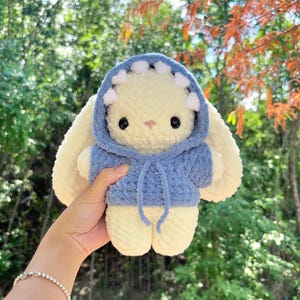 May include: A handmade, plush bunny toy with a light yellow body and large, floppy ears. The bunny wears a blue hoodie with white accents. The toy is soft and cuddly, perfect for children.