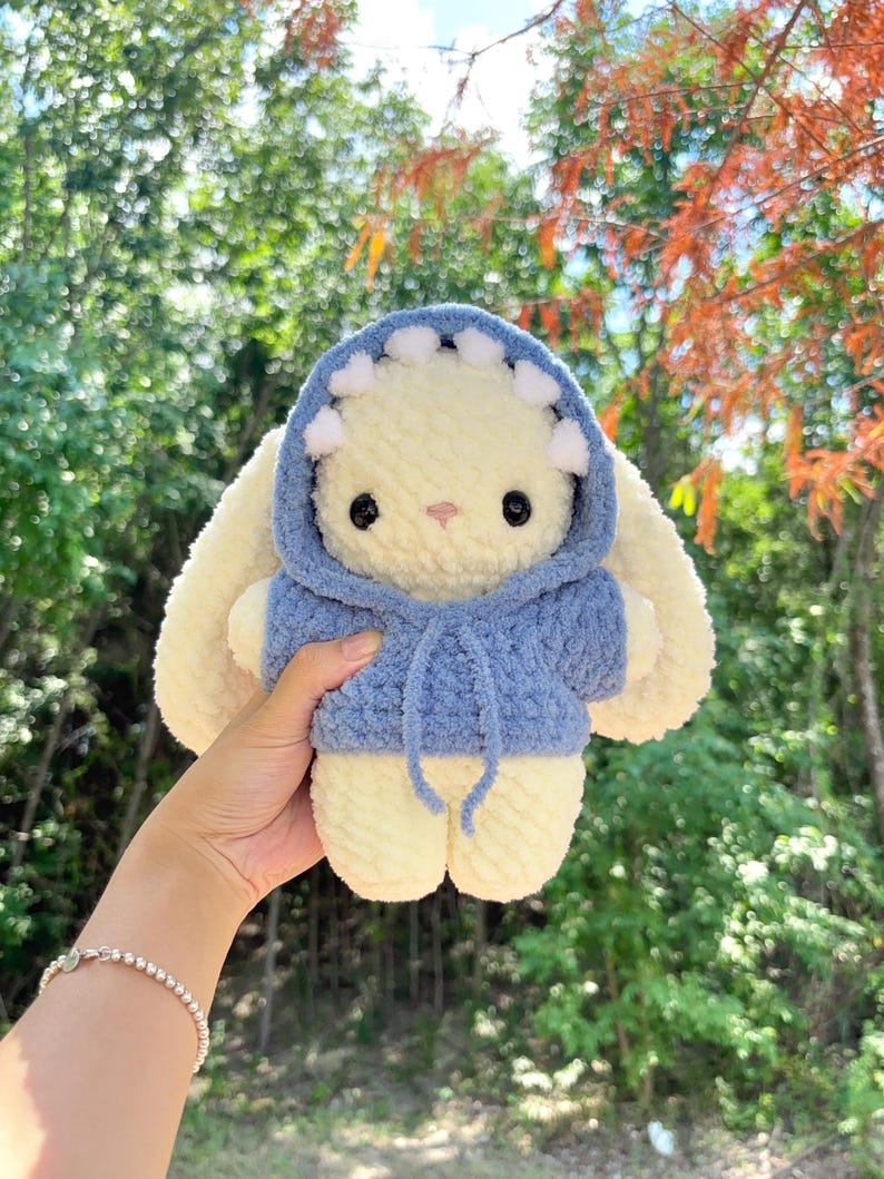 May include: A handmade, plush bunny toy with a light yellow body and large, floppy ears. The bunny wears a blue hoodie with white accents. The toy is soft and cuddly, perfect for children.
