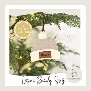 Peut inclure: Ornement de Noël en bois en forme de bonnet tricoté, avec un pompon et un bord crème. Le bonnet est beige avec une étiquette en cuir marron sur laquelle on peut lire "Hailey". L'ornement est suspendu à un cordon en cuir marron. Le texte "Laser Ready Svg" est écrit en cursive en bas.