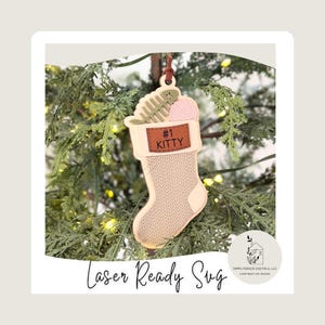 Könnte beinhalten: Ein beiger Weihnachtsstrumpf-Ornament mit einem braunen Lederaufnäher mit der Aufschrift "#1 KITTY". Der Schmuck ist mit einem Fischskelett, einem Knäuel rosa Garn und einer braunen Lederschlaufe zum Aufhängen verziert. Der Schmuck hängt an einem Weihnachtsbaum.