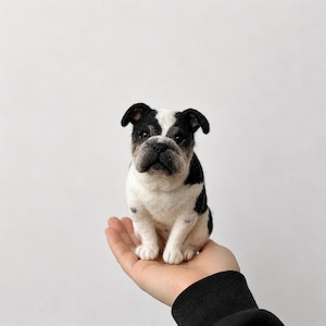 Può includere: Una statuetta di bulldog realizzata in lana infeltrita ad ago, caratterizzata da un manto bianco e nero dall'aspetto testurizzato. La piccola scultura raffigura un cane seduto con il muso rugoso, occhi scuri e orecchie pendenti, lavorata a mano.