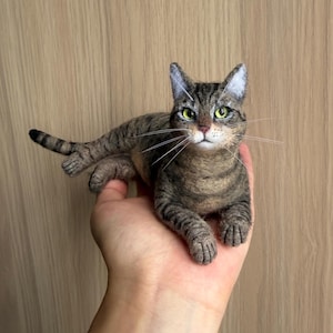 Peut inclure: Une figurine de chat tigré brun en laine feutrée avec des rayures sombres, des yeux vert vif et de longues moustaches blanches. Le chat est représenté en position de repos avec la queue étendue, montrant une texture de fourrure détaillée.