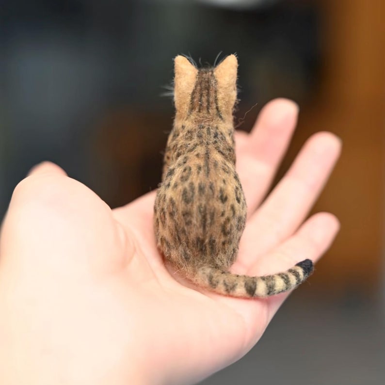 May include: A miniature felted cat figurine, viewed from the back, showcasing a spotted brown and tan coat with dark stripes. The cat's tail is striped and curled. The figurine is held in an open hand, highlighting its small size and detailed craftsmanship.