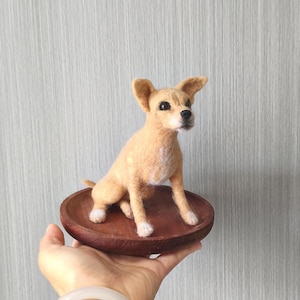 May include: A needle-felted tan dog figurine with upright ears, dark eyes, and a black nose, featuring white patches on the chest and paws. The small canine sculpture is positioned standing on a circular, smooth, dark brown wooden base.