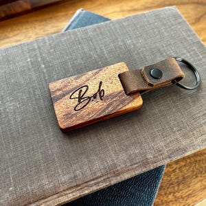 May include: A wooden keyring with a leather strap and a metal ring. The wood is a dark brown colour and has a natural grain pattern. The leather strap is a light brown colour and has a dark brown stitch. The metal ring is silver. The keyring is engraved with the name "Bob".