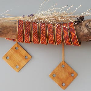 Può includere: Una fascia di tessuto intrecciato con un motivo geometrico ripetitivo in arancione, rosso e blu scuro, avvolta attorno a un pezzo di legno galleggiante. Due tavolette di legno quadrate con quattro fori circolari pendono da fili arancioni.