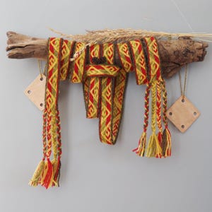 Può includere: Una cintura tessuta con un motivo geometrico in rosso, giallo e verde scuro, drappeggiata su un pezzo di legno galleggiante. La cintura termina con nappe intrecciate e fili sciolti. Due etichette in legno a forma di rombo pendono dal legno.