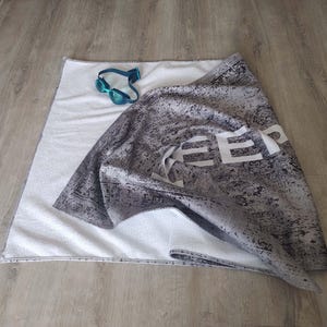 May include: A gray beach towel with white text that says "Keep Calm and Beach On". The towel is folded and has a white terry cloth backing.