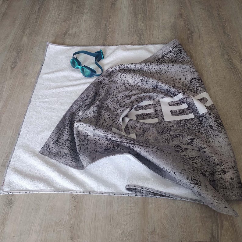 May include: A gray beach towel with white text that says "Keep Calm and Beach On". The towel is folded and has a white terry cloth backing.
