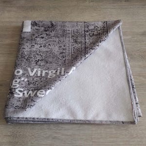 May include: A gray and white beach towel with a black and white patterned design. The towel has the text "Off-White c/o Virgil Abloh" and "Sweatshirt" printed on it.