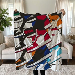 May include: A white fleece blanket with a colorful print of sneakers. The print features a variety of colors, including red, blue, black, white, and yellow. The sneakers are arranged in a random pattern, creating a dynamic and eye-catching design.