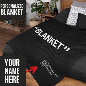 以下が含まれることがあります： 白い文字で「BLANKET」と「Blanket c/o Your Name Here BLANKET Beaverton, Oregon USA Est. 2017」と書かれた黒いフリースの毛布。毛布は枕と木製のヘッドボードのあるベッドの上に置かれています。