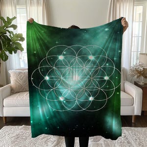 May include: A green and teal blanket with a geometric design. The blanket features a pattern of interconnected circles in a silver colour, set against a backdrop of a starry, nebula-like design. The blanket is held up by a person.