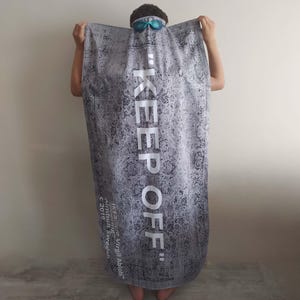 May include: A gray and white beach towel with a black and white patterned design. The towel has the text "KEEP OFF" in white letters. The text "IKEA x Off-White&trade; Virgil Abloh&trade;" is printed in black letters on the bottom of the towel.