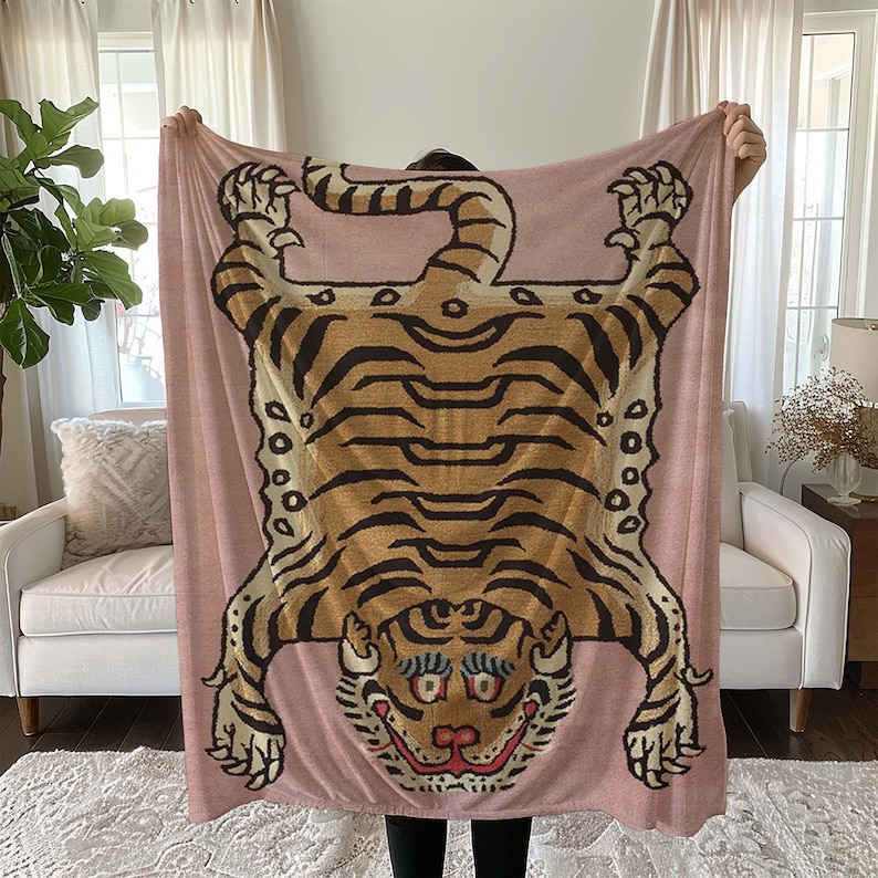 May include: A pink throw blanket featuring a tiger design. The tiger is brown and black with a pink nose and white eyes. The blanket is soft and cosy.
