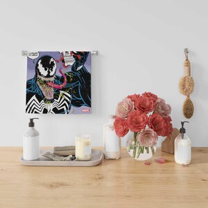 May include: A bathroom vanity with a white towel featuring a comic book image of Venom, a black and red superhero character with a large mouth and sharp teeth. The towel is hanging on a chrome towel rack. There are two clear glass bottles with white pumps, a white candle, and a vase of pink flowers on the vanity.