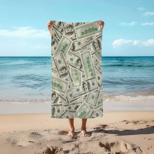 Könnte beinhalten: Ein Strandtuch mit einem Muster aus US-Dollar-Scheinen. Das Handtuch wird von einer Person gehalten, die auf einem Sandstrand steht.