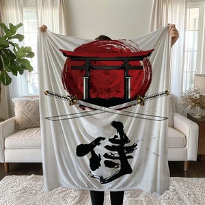 May include: White fleece blanket featuring a red and black Japanese-style gate, two swords crossed behind it, and a black kanji character in the bottom centre. The gate is set against a red circle with a white outline.
