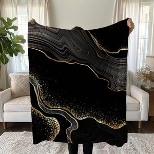 May include: A black blanket with a gold and gray marble design. The blanket features flowing lines and gold accents, creating a luxurious and elegant aesthetic. The design is reminiscent of natural stone.