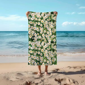 Op de afbeelding: Een witte strandhanddoek met een patroon van witte madeliefjes op een groene achtergrond. De handdoek wordt vastgehouden door een persoon op een strand.