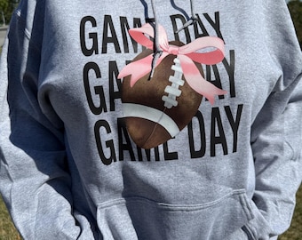 Może przedstawiać: Szara bluza z kapturem z czarnym napisem "GAME DAY". Brązowa piłka do futbolu amerykańskiego z białymi sznurowadłami i różową kokardą pośrodku. Bluza ma przednią kieszeń i kaptur ze sznurkiem.