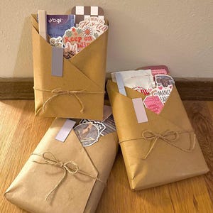 Può includere: Tre pacchi di carta marrone legati con fiocchi di spago. Ogni pacco, piegato come una busta, contiene adesivi e piccoli oggetti. Uno ha un adesivo con scritto "Keep on Doin", un altro "Romance Reads".