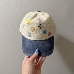 May include: A beige baseball cap with a denim blue brim. The cap is decorated with various smiley face and daisy patches in yellow, pink, blue, and white. The cap is held by a person.
