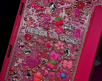 May include: Pink iPad case with a glittery, resin-filled design. The case is covered in pink, white, and silver glitter, and features various charms, including hearts, stars, bows, and a teddy bear. The text "Nessa Da Costa" is written in silver glitter on the case.